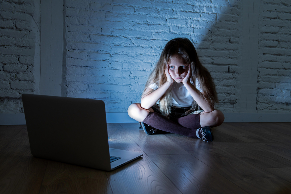 A young girl sits in a dark room with a computer on the floor.