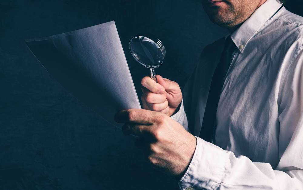 Close dark photo of man in suit holding magnifying glass up to paper